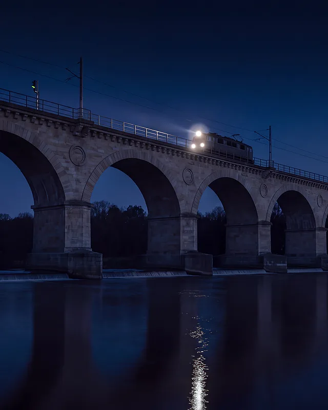 Kamienny wiadukt kolejowy nad Bobrem w Bolesławcu o zmierzchu; po wiadukcie jedzie pociąg z zapalonymi reflektorami, a ich światło odbija się w rzece.