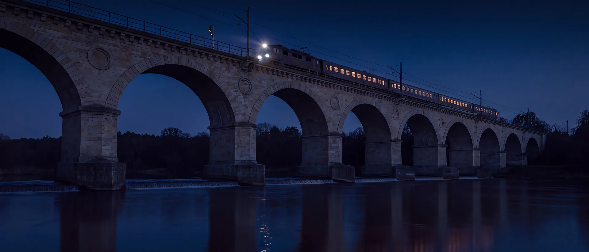 Kamienny wiadukt kolejowy nad Bobrem w Bolesławcu o zmierzchu; po wiadukcie jedzie pociąg z zapalonymi reflektorami, a ich światło odbija się w rzece.