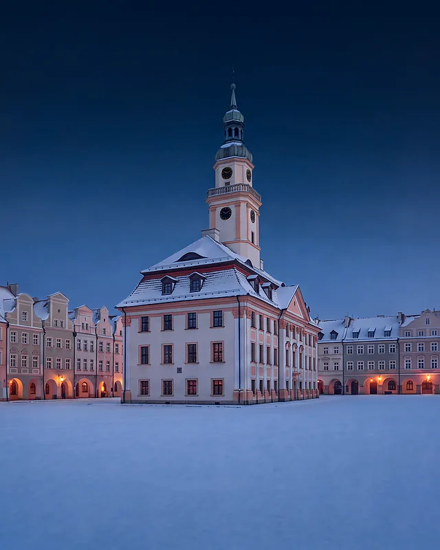 Ratusz na placu Ratuszowym w Jeleniej Górze zimą o zmierzchu: zaśnieżony plac, wieża ratusza i kamienice z podcieniami oświetlonymi latarniami