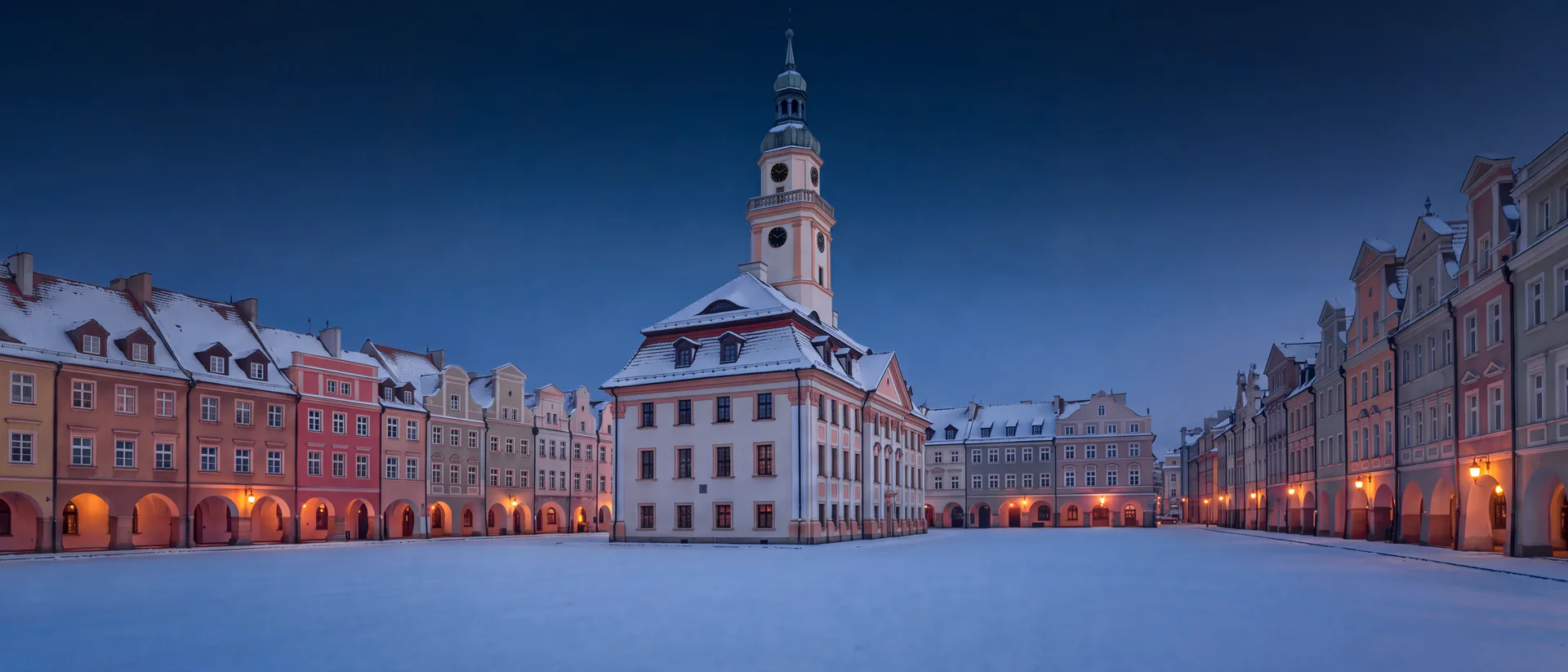 Ratusz na placu Ratuszowym w Jeleniej Górze zimą o zmierzchu: zaśnieżony plac, wieża ratusza i kamienice z podcieniami oświetlonymi latarniami
