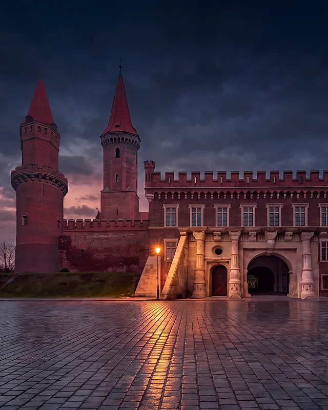 Zamek Piastowski w Legnicy o zmierzchu po deszczu: renesansowa brama w ceglanym skrzydle z blankami, nad nią wieża św. Piotra, z lewej wieża św. Jadwigi; światło latarni odbija się w mokrym bruku placu