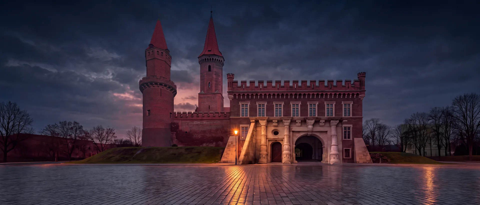 Zamek Piastowski w Legnicy o zmierzchu po deszczu: renesansowa brama w ceglanym skrzydle z blankami, nad nią wieża św. Piotra, z lewej wieża św. Jadwigi; światło latarni odbija się w mokrym bruku placu