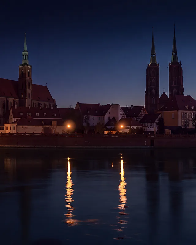 Ostrów Tumski we Wrocławiu nocą: Most Tumski, kolegiata Świętego Krzyża i dwie wieże katedry nad Odrą; światło latarni gazowych odbija się w rzece długimi smugami.