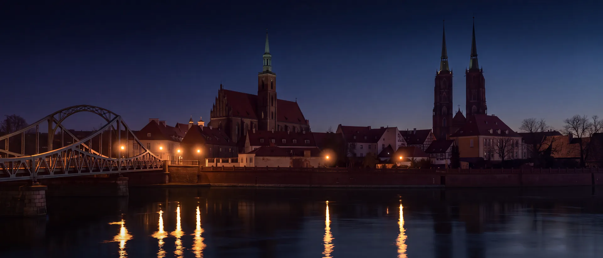 Ostrów Tumski we Wrocławiu nocą: Most Tumski, kolegiata Świętego Krzyża i dwie wieże katedry nad Odrą; światło latarni gazowych odbija się w rzece długimi smugami.