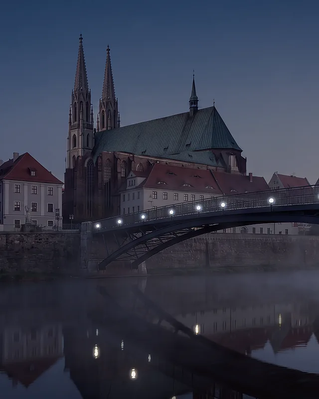 Most Staromiejski na Nysie o świcie: stalowy łuk kładki nad zamgloną rzeką, na drugim brzegu kamienice Görlitz i kościół św. Piotra i Pawła z dwiema wieżami; na moście palą się latarnie.
