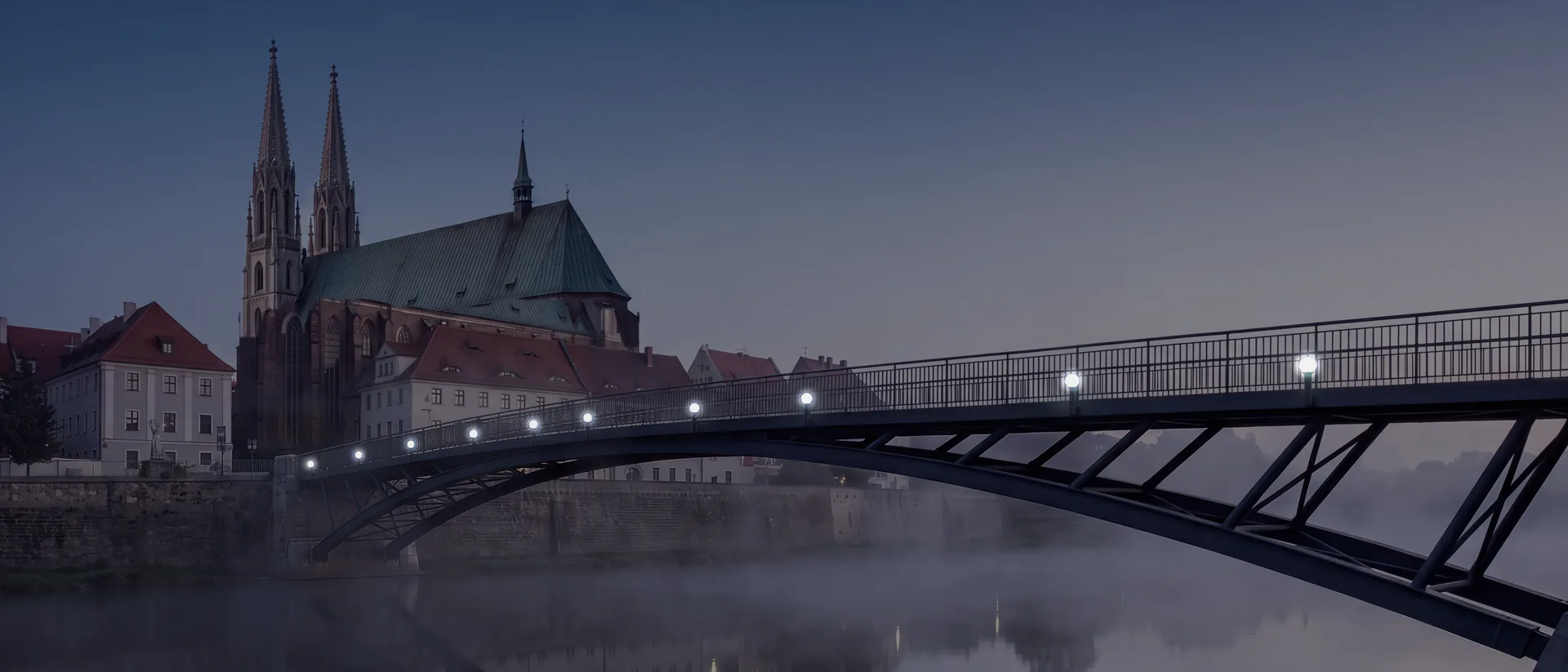 Most Staromiejski na Nysie o świcie: stalowy łuk kładki nad zamgloną rzeką, na drugim brzegu kamienice Görlitz i kościół św. Piotra i Pawła z dwiema wieżami; na moście palą się latarnie.